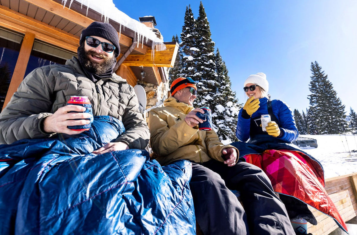 three friends with rumpl blankets and beers in rumpl beer blankets sharing an après-ski moment outside a lodge