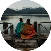 a man and woman sitting at a pier with the baja fade down puffy blanket around their shoulders, the view of a lake and the mountains in the background.